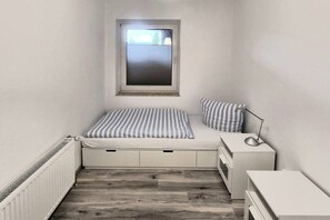 A small, compact bedroom with a single bed, a bedside table, a lamp, and a window with blinds. - Holiday apartment with 1 bedroom (Norddeich)