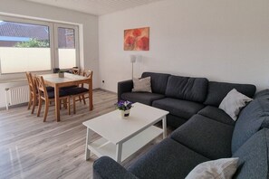 A modern living room with a grey sofa, a white coffee table, and a dining area with wooden chairs and a table. - Holiday apartment with 1 bedroom (Norddeich)