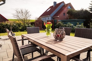 Een terras buiten met een houten tafel en rieten stoelen, een vaas met rode bloemen, en een bakstenen huis op de achtergrond.