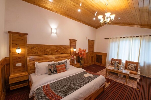 Una habitación de hotel con cabecera de madera, cama con ropa de cama estampada, zona de estar con sofá y sillones, y un candelabro.