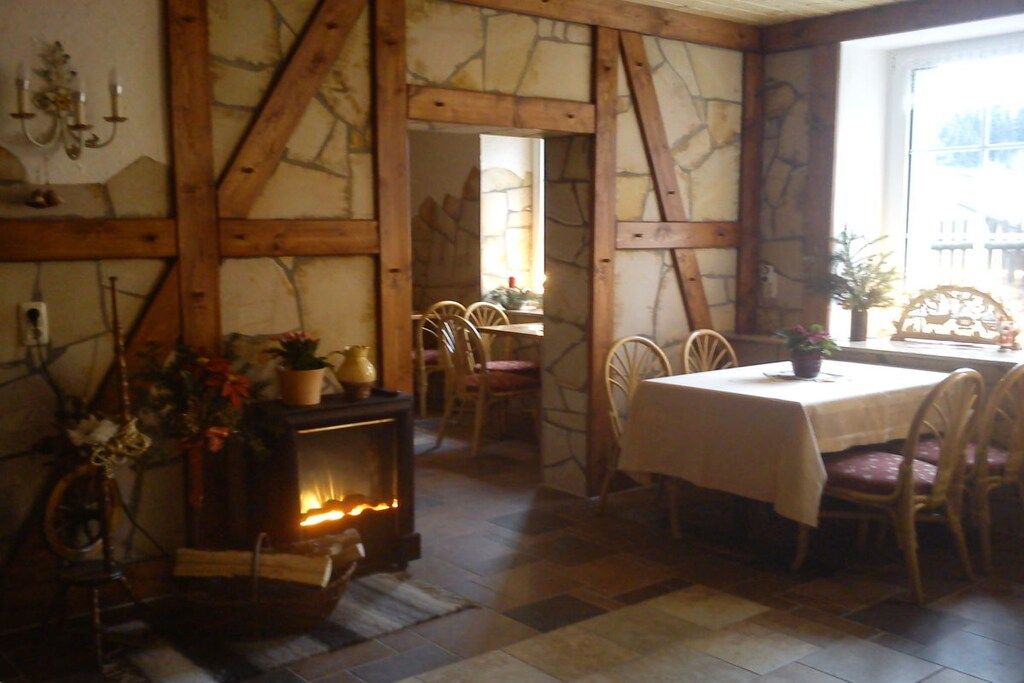 Un interior rústico con vigas de madera a la vista, pared de piedra, chimenea y zona de comedor con sillas y mesa.
