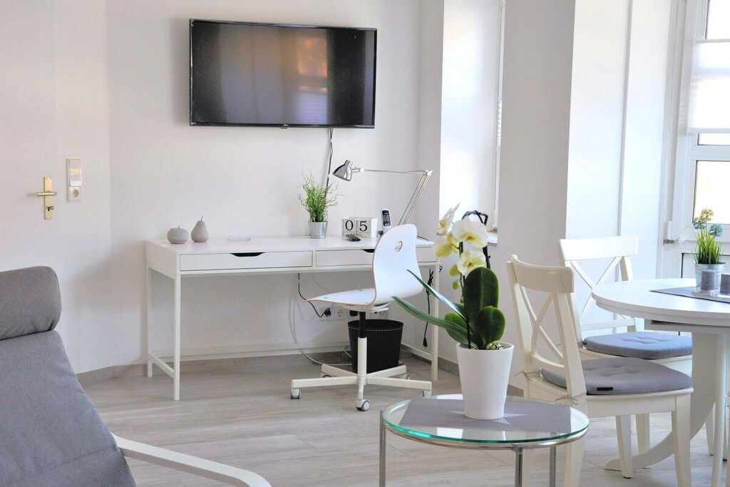 Een moderne leefruimte met een flatscreen-tv aan de muur, een wit bureau met een computer, een stoel en een kleine potplant.