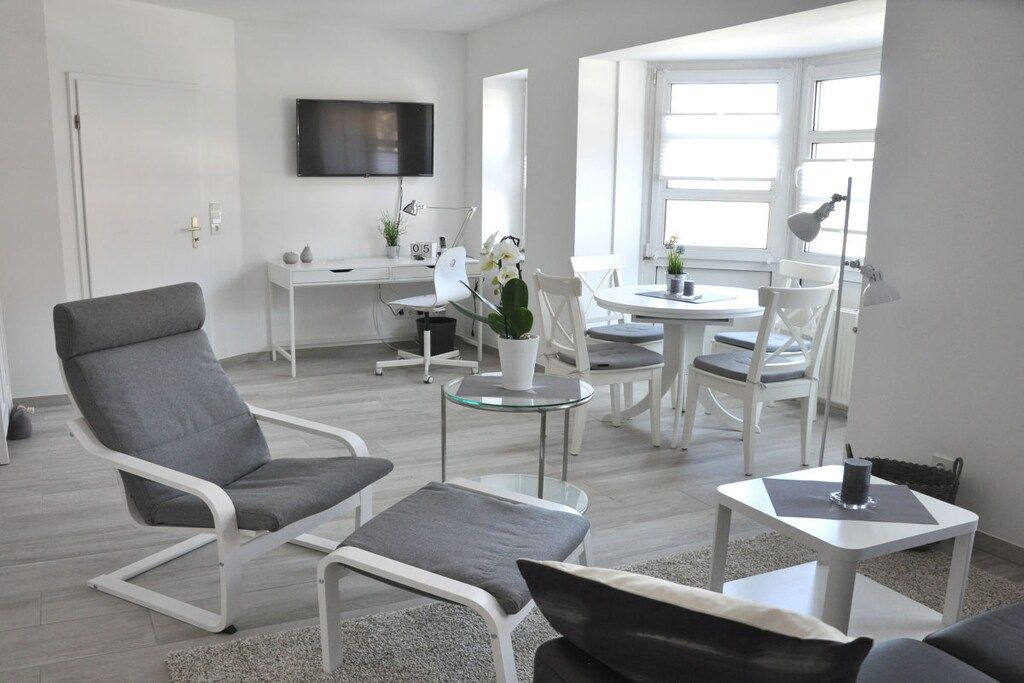 A modern living room with a grey sofa, a white armchair, a glass coffee table, and a dining area with a round table and chairs.