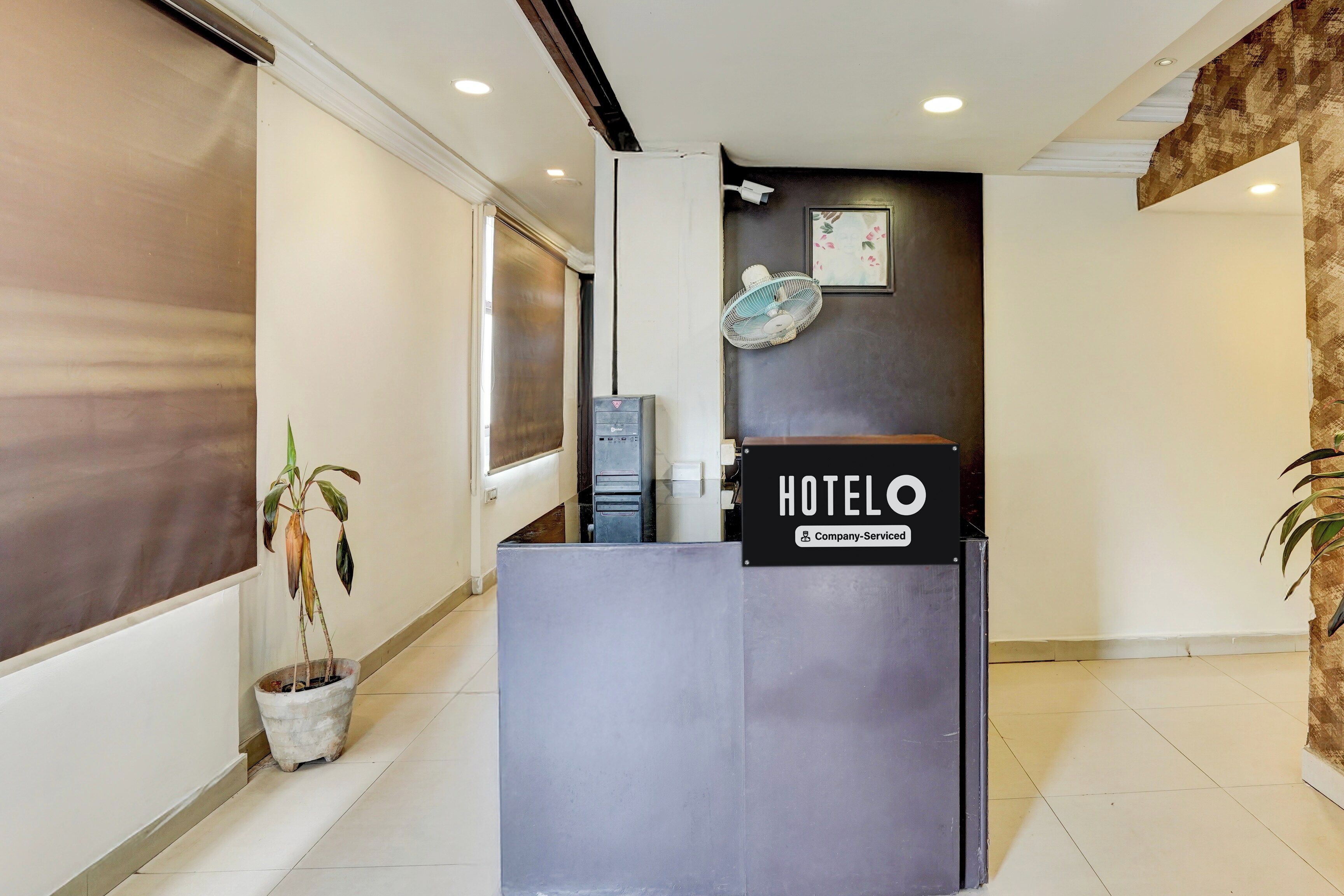 A hotel reception desk with a sign indicating it is company-serviced. There is a potted plant to the left and a wall-mounted fan to the right.