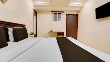 A hotel room with a bed, bedside table, a small desk, a chair, a potted plant, and a TV mounted on the wall.