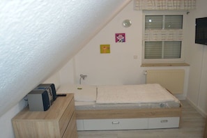 Une petite chambre avec un lit simple, un plafond en pente, un radiateur, une fenêtre avec des rideaux, un bureau avec une imprimante et une lampe fixée au mur.