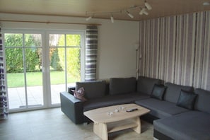 A modern living room with a sectional sofa, a wooden coffee table, and a sliding glass door leading to a garden. - Mühlenblick 32 - Wohlgefahrt (Wittmund-Carolinensiel)