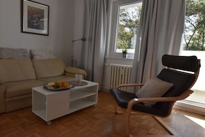 A living room with a sofa, a coffee table with a tray of fruit, a chair, and a window with curtains. - Holiday apartment in Kiel with swimming pool (Kiel)