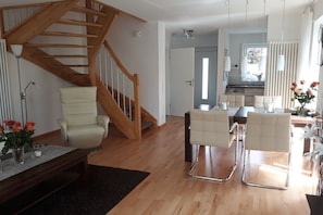 A modern living room with a wooden staircase, a dining area with white chairs, and a kitchen in the background. - Holiday home with 2 bedrooms (Wangerland-Hooksiel)