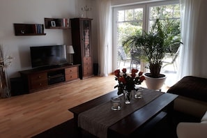 Ett vardagsrum med en tv, ett träinrett mediaskåp, en krukväxt och ett fönster med utsikt över grönområden.