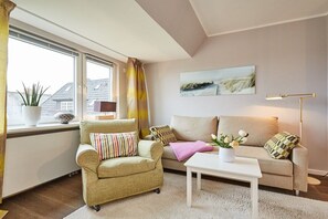 Ein Wohnzimmer mit Sofa, Sessel und Couchtisch. Ein Fenster mit Vorhängen, eine Pflanze auf der Fensterbank und ein Bild an der Wand.