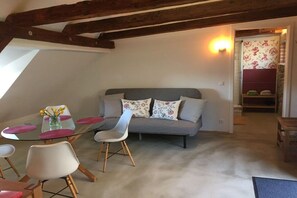 A cozy living area with a grey sofa, white and floral cushions, a glass table with pink placemats, and a wooden chair. - Holiday apartment with 1 bedroom (Meißen)