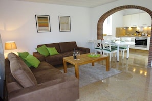 Ein Wohnzimmer mit einem braunen Sofa, grünen Kissen, einem Holztisch und einem Essbereich mit weißem Tisch und Stühlen.