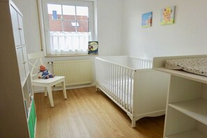 A room with a crib, a chair, and a window with a view of buildings. - FH North Sea Feeling LD 113a (Hooksiel)