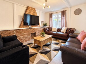 A living room with a brick fireplace, a flat-screen TV mounted above it, a brown leather sofa, a wooden coffee table, and a patterned rug. - Spacious 3BDR Home with Free Parking Sleeps 6 (Basildon)