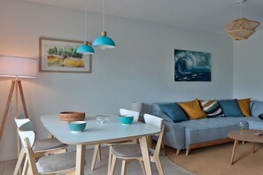 A modern living room with a white table, blue chairs, a blue sofa with patterned cushions, and a painting of a wave on the wall. - Holiday apartment 5 - Flounder (Börgerende)