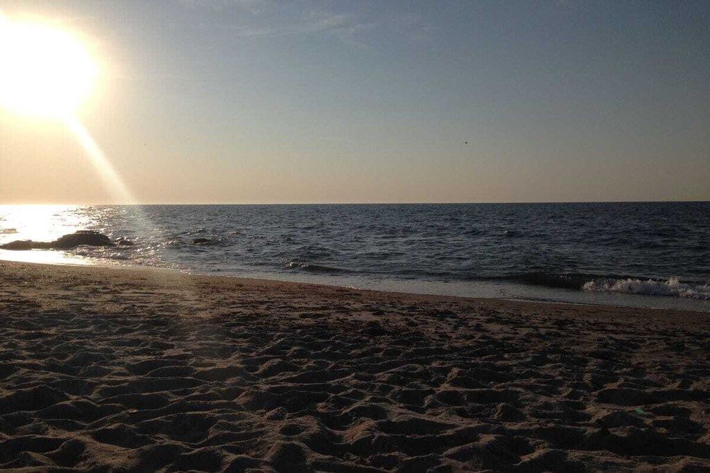Una playa con arena y agua, y el sol visible en el cielo.
