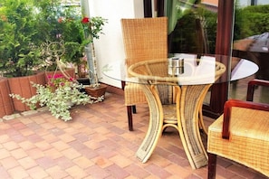 A wicker table and chairs set on a brick patio with a glass top, surrounded by potted plants and a view of greenery. - Holiday apartment with 2 bedrooms (Karlshagen)