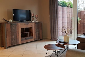 A living room with a TV on a wooden cabinet, two small tables, and a window with a view of a brick wall and greenery. - Holiday apartment with 2 bedrooms (Karlshagen)