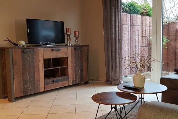 A living room with a TV on a wooden cabinet, two small tables, and a window with a view of a brick wall and greenery. - Holiday apartment with 2 bedrooms (Karlshagen)
