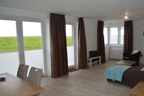 A room with a dining table, chairs, a sofa, a TV, and large windows with curtains. - New building with dike view & south-facing terrace (Juist)