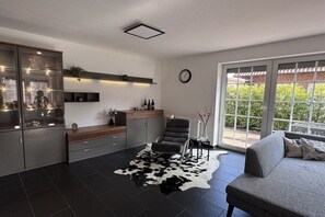A modern living room with a sofa, a glass cabinet, a dining area, and a large window. - Holiday home with 3 bedrooms (Wangerland)