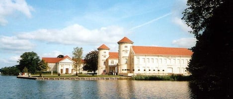 A large building with a red roof and multiple towers, situated by a calm body of water.