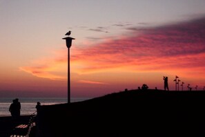 Silhouetten van mensen op een strand bij zonsondergang, met een lantaarnpaal en windmolens op de achtergrond.