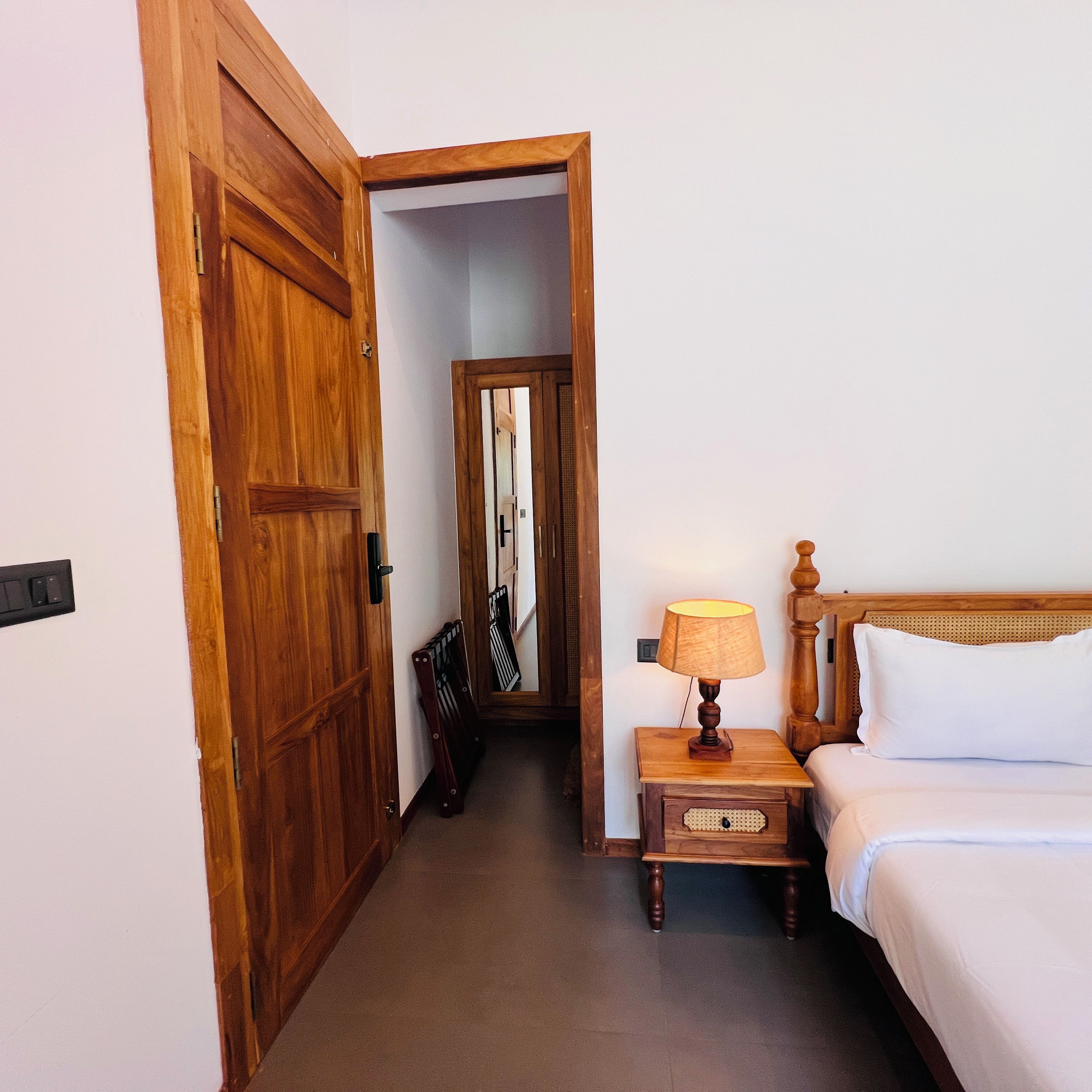 A hotel room with a wooden door, a bedside table with a lamp, and a bed with white bedding.