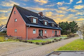 A red brick building with a steeply pitched roof, a front entrance with steps, and a paved driveway. - Holiday apartment with 2 bedrooms (Wenningstedt)