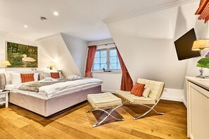 Un dormitorio con una cama, un sillón chaise, un televisor y un ventanal con cortinas.