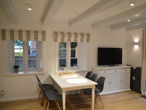 A dining area with a white table, black chairs, a TV on a cabinet, and windows with curtains. - 1 Aruba at Haus Boldix (Wyk auf Foehr)