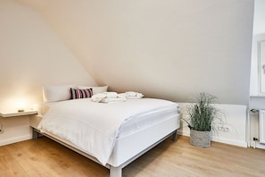 A bed with white linens and a striped pillow, a bedside table with a lamp, and a potted plant. - Holiday apartment with 2 bedrooms (Wenningstedt)