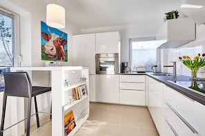 A modern kitchen with white cabinets, a built-in oven, a coffee maker, and a bar area with a stool. - Holiday apartment in Sylt with 3 bedrooms (Westerland)