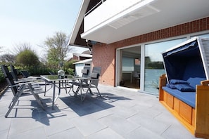 Une terrasse extérieure avec une table et des chaises, un banc en bois avec des coussins et une porte vitrée donnant sur une pièce.