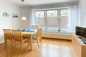 A room with a dining table, four chairs, a television, and a large window with a view of a brick wall and greenery. - Holiday apartment in Juist with swimming pool (Juist)