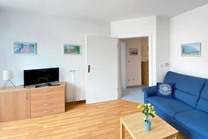 A living room with a television on a wooden cabinet, a blue sofa, and a small table with a vase of flowers. - Holiday apartment in Juist with swimming pool (Juist)