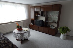 A living room with a television, a cabinet with glassware, a round table with a lace tablecloth, a potted plant, and a window with curtains. - Holiday apartment with 1 bedroom (Ebersbach-Neugersdorf)