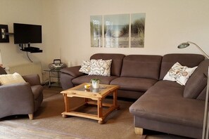 A living room with a brown sofa, a glass coffee table, and a television on a stand. - Holiday home in Hooksiel with swimming pool (Hooksiel)