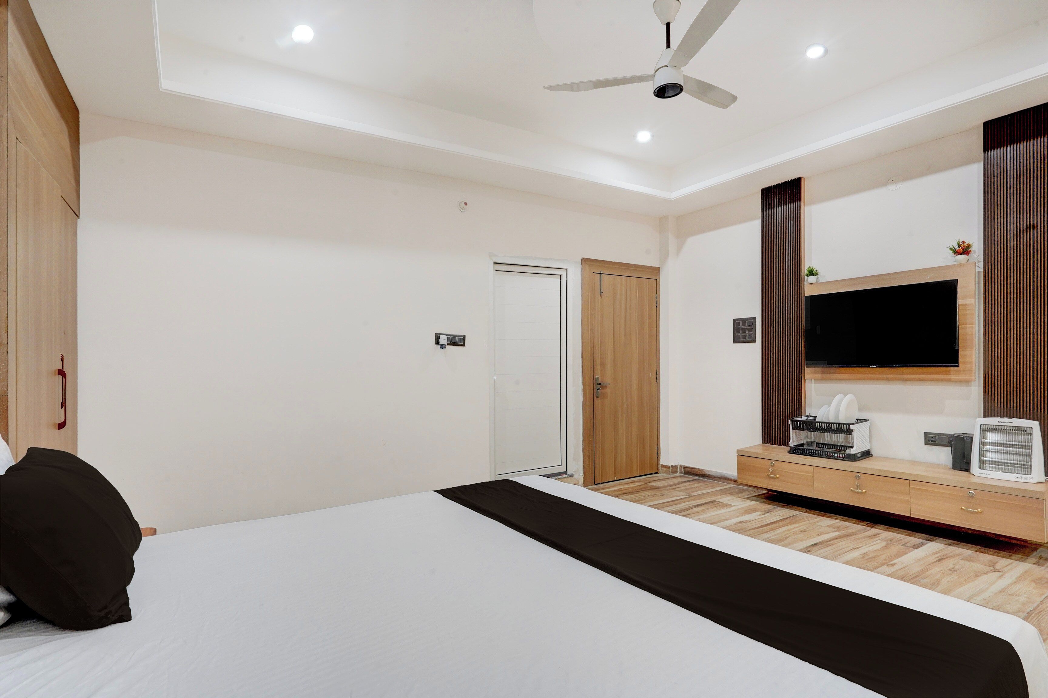 A hotel room with a bed, a ceiling fan, a TV, a small table with a heater, and a wooden cabinet.