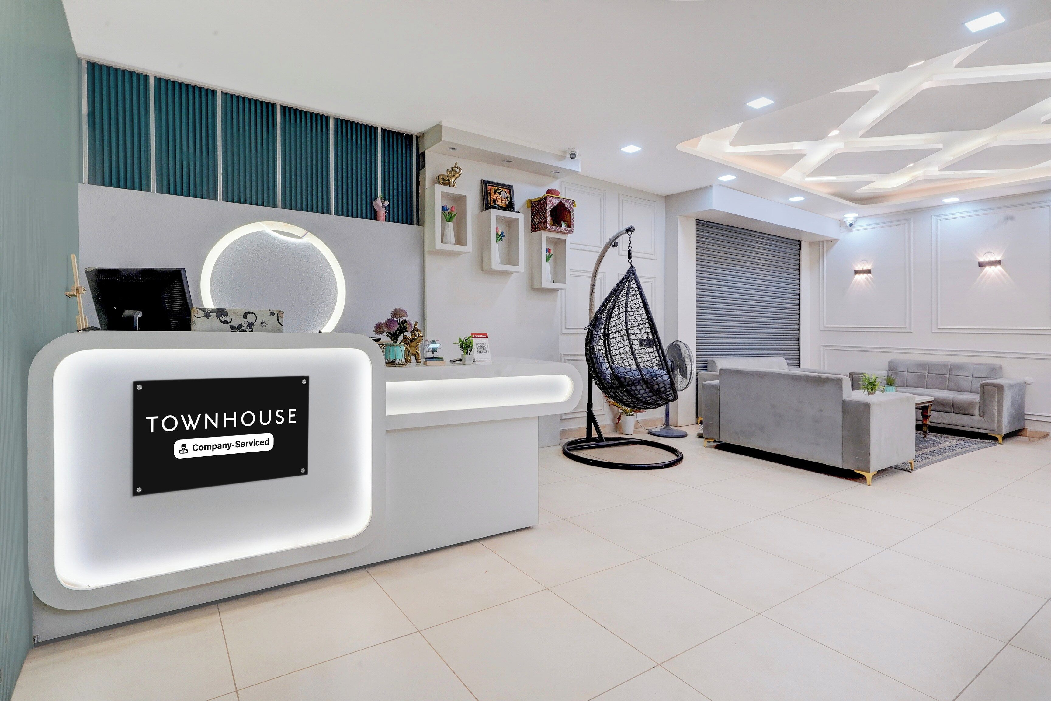 A modern reception desk with a 'TOOWNHOUSE Company-Serviced' sign, a hanging chair, and a grey sofa in a well-lit lobby area.