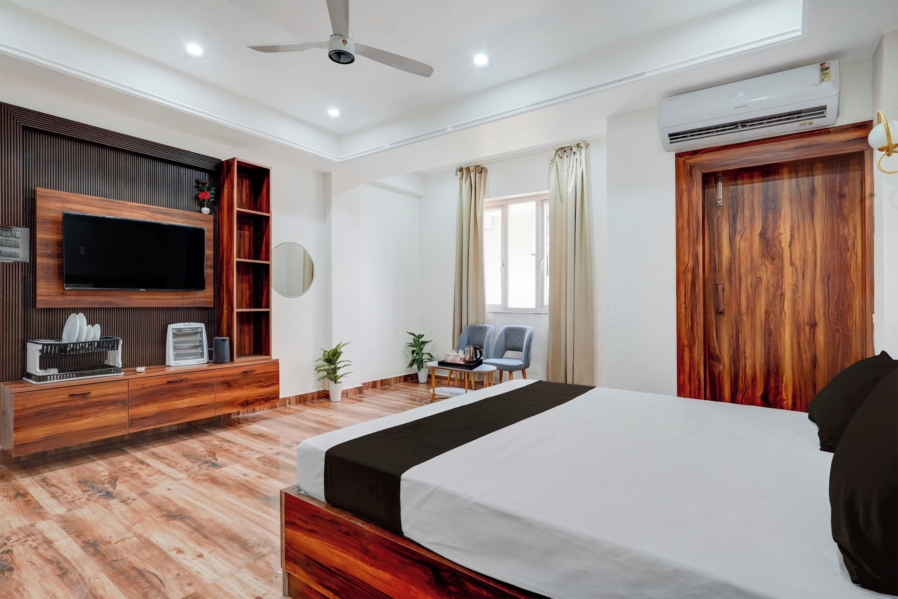 A hotel room with a bed, a TV mounted on the wall, a wooden wardrobe, a small table with chairs, and an air conditioning unit.