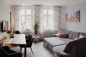 A modern living room with a grey sofa, a dining table with chairs, a potted plant, and a cityscape painting on the wall. - Apartment in Quedlinburg with swimming pool (Quedlinburg)