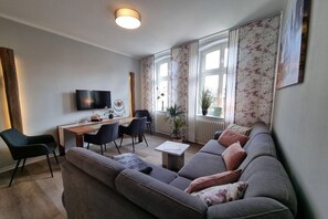 En moderne stue med sofa, spisebord med stole, fladskærms-tv og store vinduer med blomstrede gardiner.