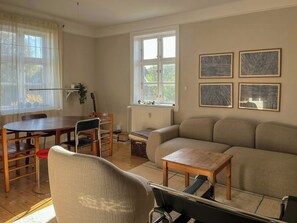 A living room with a sofa, a dining table with chairs, a radiator, and a window with curtains. - 1930s flat with a renovated garden in Valby (København)