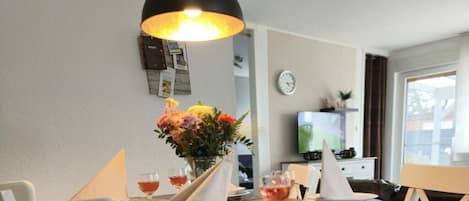 A dining area with a table set for two, a vase with flowers, and a television in the background.
