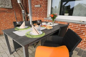 An outdoor dining area with a brick wall, a window, and a table set with plates, glasses, and a vase of flowers. - Bungalow with 1 bedroom (Dornum - Dornumersiel)
