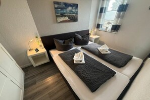 A hotel room with two beds, a nightstand with a lamp, a painting on the wall, and a window with curtains. - Bungalow with 1 bedroom (Dornum - Dornumersiel)
