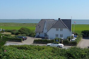 A white building with a steeply pitched roof, situated on a green landscape with a view of the sea. - Holiday apartment with 1 bedroom (Rantum)