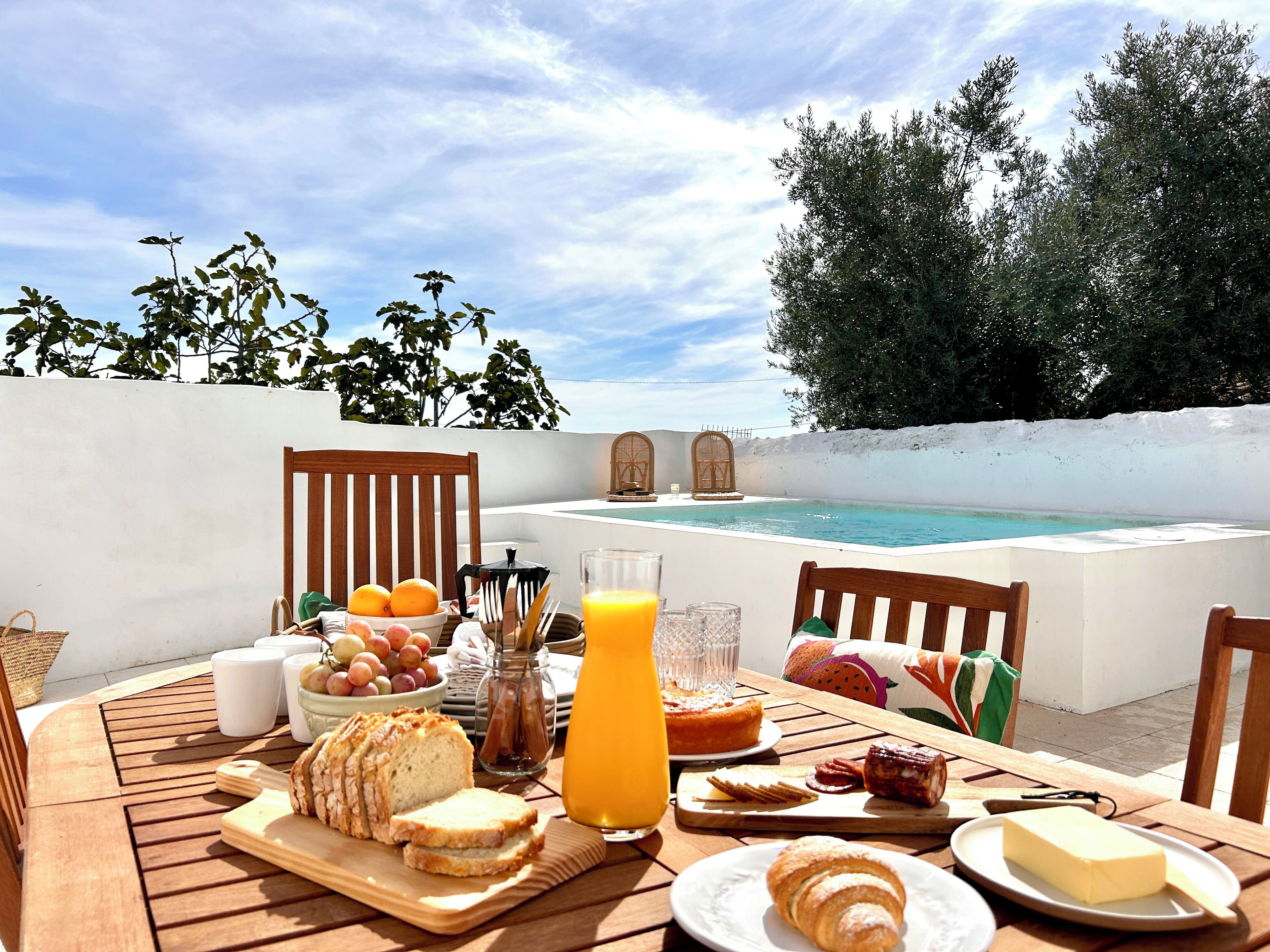 A breakfast table with bread, fruit, and a glass of orange juice, overlooking a swimming pool and trees.